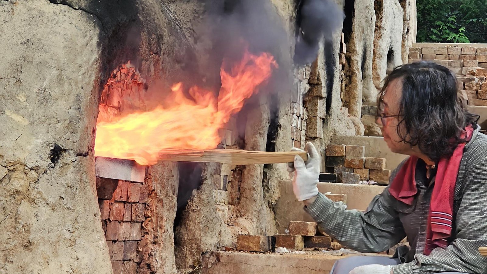 Shin Hyun Moon, accroupi, alimente en bois le foyer d’un four traditionnel d’où sortent de hautes flammes