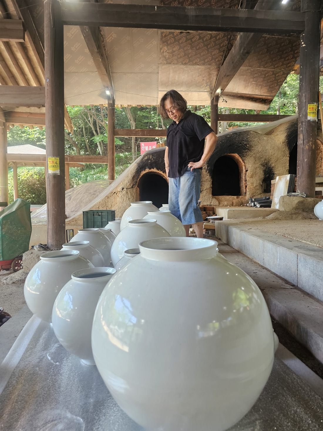 Des dizaines de moon jars blancs émaillés posés au sol sous l’abri d’un four à bois ; Shin Hyun Moon se tient au fond