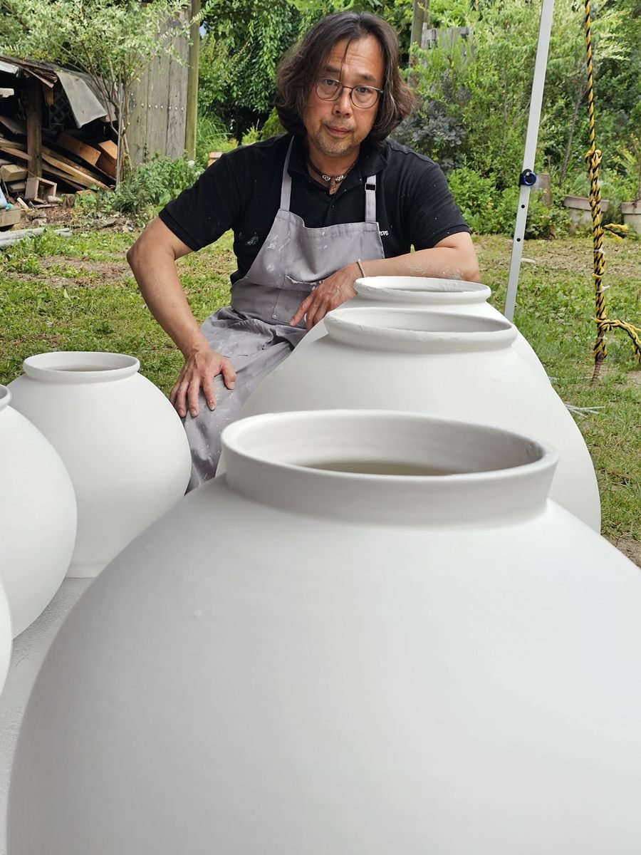 Shin Hyun Moon, souriant, appuyé sur des moon jars crus disposés dans l’herbe